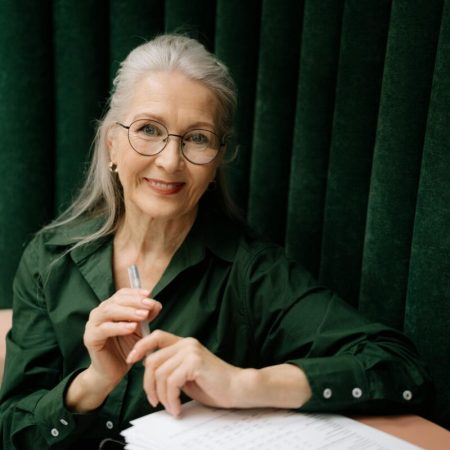 Elegant senior woman with eyeglasses smiling while holding a pen and reviewing documents indoors.