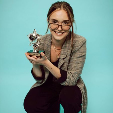 Woman in formal attire with eyeglasses holding a Best Teacher trophy, smiling against a green background.